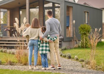 Happy family. View from back of man woman with child and girl standing together hugging and admiring their cozy home on fine autumn day